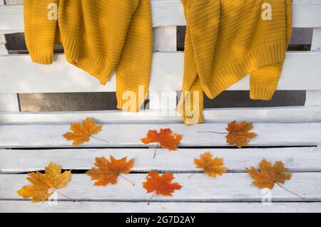 Deux chandails tricotés de couleur moutarde sont suspendus à l'arrière d'un banc en bois blanc. Des feuilles d'érable orange-rouge se trouvent à proximité. Atmosphère d'automne Banque D'Images