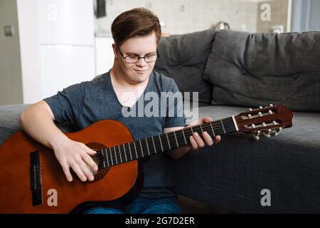 Jeune homme avec le syndrome de Down jouant de la guitare tout en étant assis sur le sol à la maison Banque D'Images