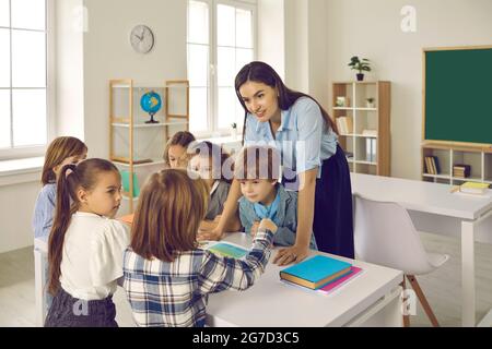 Souriant amical jeune femme enseignant caucasien aide et explique le matériel scolaire aux enfants. Banque D'Images