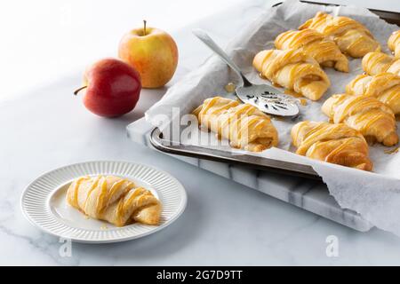 Rouleau croissant rempli de pomme sur une petite plaque avec une plaque de cuisson de plus à l'arrière, contre une fenêtre lumineuse et ensoleillée. Banque D'Images