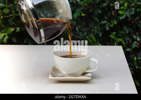 Versez le café de la casserole à la tasse qui se trouve sur une table blanche, avec des feuilles vertes floues sur l'arrière-plan. Banque D'Images