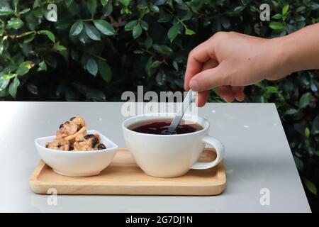 Tenir la cuillère à la main en remuant le café sur le plateau en bois et il y a des biscuits sur le côté. La table est située dans le jardin avec un fond de feuilles vertes. Copier l'espace. Banque D'Images