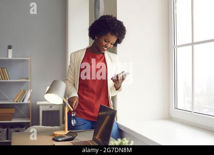 Bonne jeune femme d'affaires utilisant un téléphone portable debout près de la fenêtre dans son bureau Banque D'Images