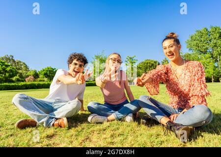 Trois jeunes étudiants meilleurs amis assis sur l'herbe dans le parc de la ville montrant le signe de victoire avec deux doigts regardant la caméra. Concept d'unité et de solidité Banque D'Images