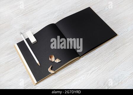 Ouvre un carnet d'esquisses noir vierge, un crayon et une gomme sur fond de table en bois clair. Banque D'Images
