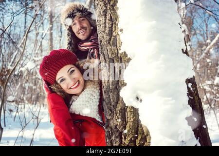 Couple espiègle se cachant derrière un tronc d'arbre dans la neige en regardant dans la caméra Banque D'Images