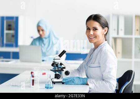 Scientifique souriant en gants de latex regardant la caméra près du microscope et de l'équipement Banque D'Images