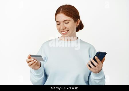 Image d'une femme souriante aux cheveux rouges et aux taches de rousseur, utilisant une application, un téléphone mobile et une carte de crédit avec un visage heureux et détendu, un paiement facile en ligne, des achats Banque D'Images