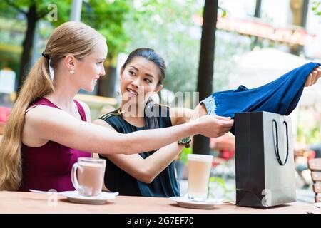 Jolie femme asiatique montrant à un ami son achat d'un vêtement bleu alors qu'ils s'assoient à une table de restaurant en appréciant des rafraîchissements Banque D'Images