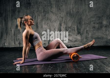 Une jeune femme athlétique musclée avec un corps magnifique, portant des vêtements de sport avec un masseur à rouleau en mousse assis sur un tapis de yoga. Banque D'Images