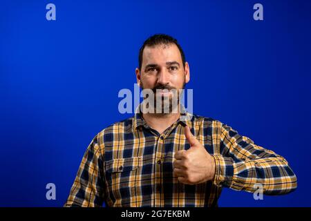 Un bon homme qui donne les pouces vers le haut signe comme le geste des mains. Petit garçon avec barbe en chemise à carreaux bleu isolée sur fond gris studio. Négatif Banque D'Images