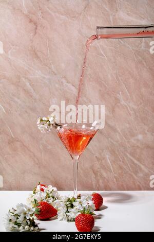 Fraise déversant un cocktail alcoolisé ou non alcoolisé d'une bouteille de verre dans un verre martini, décoré de branches de cerisier fleuris sur blanc Banque D'Images