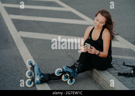 Vue de dessus de brunette active jeune femme utilise smartphone pour la communication en ligne poses sur l'asphalte vêtu de noir active port aime passe-temps favori Banque D'Images