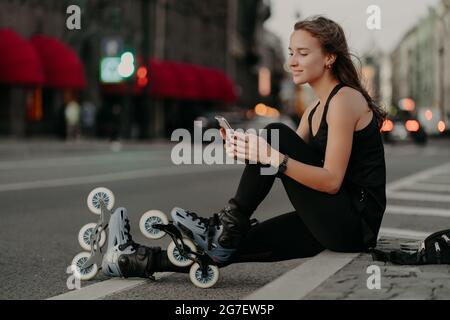 Prise de vue horizontale d'une femme sportive active vêtue de vêtements de sport porte des rollerblades focalisées sur le smartphone. Elle aime le patinage en ligne en plein air Banque D'Images