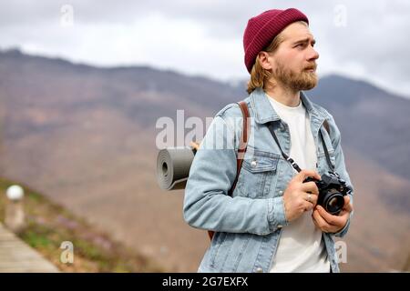 Beau caucasien homme avec sac à dos et appareil photo est en marche vers la montagne éloignée. Bearded Man ranking dans les montagnes au printemps. Voyage de voyageur Banque D'Images
