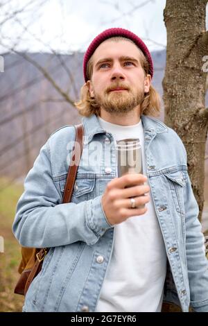 jeune beau barbu brutal voyage dans la nature sauvage avec des thermos, automne ou printemps style, voyageur en tenue décontractée ont le repos, debout à côté Banque D'Images