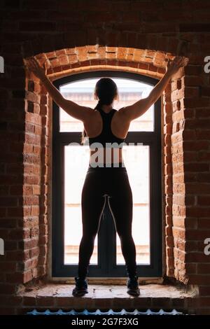 Vue arrière d'une jeune femme sportive musclée avec un corps solide et beau dans des vêtements de sport noirs gardant les mains debout sur le rebord de la fenêtre par la fenêtre. Banque D'Images