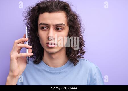 Ennuyé curly homme ont parler sur smartphone, à l'écoute de parler, jeune homme ayant le visage sérieux, dans un t-shirt décontracté, isolé sur fond violet Banque D'Images