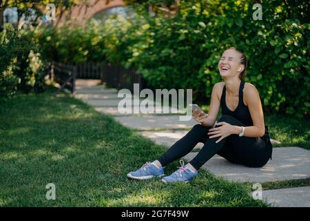 Le sportswoman débordé de joie est assis sur le chemin de rire utilise un smartphone pour écouter de la musique de la liste de lecture partager du contenu et discuter vêtu d'un vêtement actif Banque D'Images