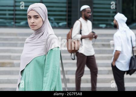 Portrait d'une jeune femme musulmane sérieuse et confiante en vêtements nationaux portant un sac à bandoulière contre ses collègues à l'extérieur Banque D'Images