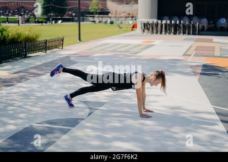 Sportive mince jeune femme fait l'exercice de planking soulève des jambes démontre sa détermination porte des vêtements de sport et des baskets trains en plein air écoute Banque D'Images