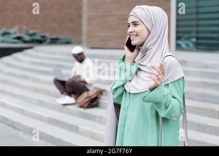 Jeune femme arabe gaie et attrayante dans le hijab se tenant contre la construction de la ville et partageant des expressions par téléphone Banque D'Images