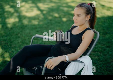 Photo en plein air d'une belle femme aux cheveux foncés habillée de vêtements de sport, boissons à emporter, café passe du temps libre dans des poses en plein air sur une chaise Banque D'Images