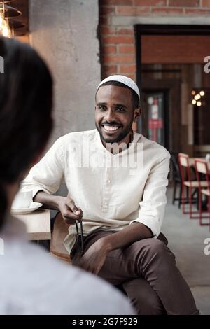 Positif jeune Noir homme en casquette kufi assis à la table dans le café loft et parlant à un ami Banque D'Images