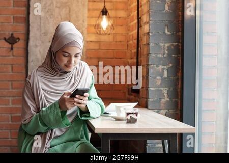 Contenu jeune femme musulmane dans hijab assis à une table haute dans le café loft et SMS sur le téléphone Banque D'Images