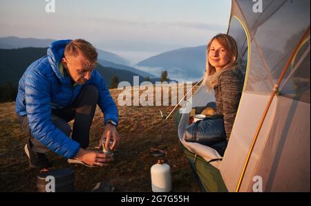 Charmante jeune femme randonneur assis à l'intérieur du camp tente et souriant pendant que l'homme prépare une boisson chaude. Les jeunes voyageurs en couple utilisent des équipements touristiques pour faire du café tout en faisant de la randonnée dans les montagnes. Banque D'Images