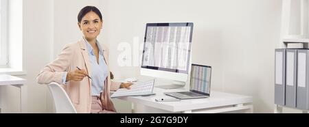 Bonne jeune femme d'affaires assise au bureau avec un ordinateur de bureau et un ordinateur portable Banque D'Images