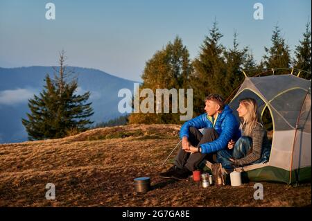 Vue latérale de couple de voyageurs mariés se reposant dans une tente sur un pré de montagne pendant la randonnée d'automne sur la toile de fond des arbres et de la colline de montagne. Matin de soleil dans le camping. Banque D'Images