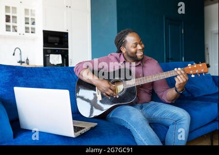 Emmenée en jouant de la guitare afro-américaine gars utilisant un ordinateur portable pour jouer, streaming en ligne, homme noir talentueux avec guitare acoustique joue devant le latop assis sur le canapé à la maison Banque D'Images