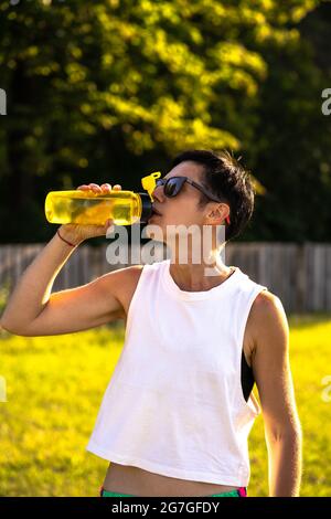 Jeune belle femme boire de l'eau de la bouteille en plastique dans un parc après l'entraînement, cheveux noirs courts, lunettes de soleil et short, sport en plein air en été, Banque D'Images