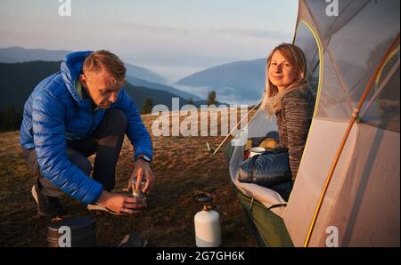 Charmante jeune femme randonneur assis à l'intérieur du camp tente et souriant pendant que l'homme prépare une boisson chaude. Les jeunes voyageurs en couple utilisent des équipements touristiques pour faire du café tout en faisant de la randonnée dans les montagnes. Banque D'Images
