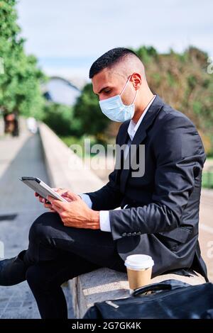 Vue latérale d'un jeune homme d'affaires concentré en costume élégant et masque de protection lisant les informations sur une tablette tout en ayant une pause avec une tasse de café à emporter Banque D'Images