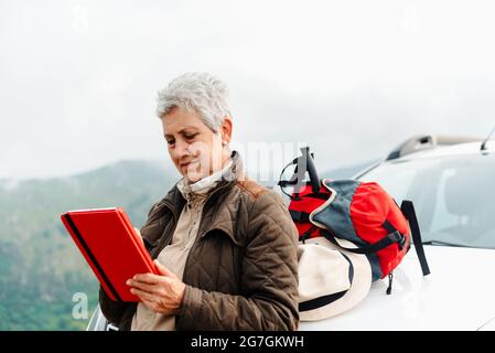 Femme voyageur âgée aux cheveux gris courts penchée sur la voiture et parcourant une tablette lors d'un voyage en campagne Banque D'Images