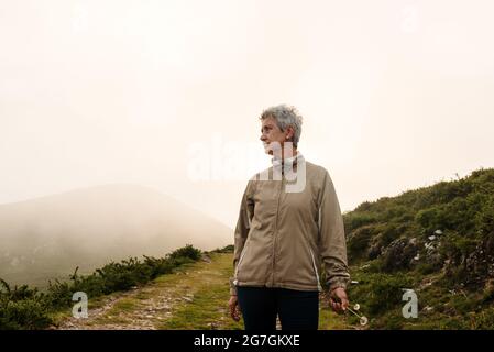 Voyageur âgé avec de courts cheveux gris qui regarde loin et marche sur le sentier près de la colline en journée dans la nature Banque D'Images