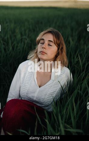 Paisible jeune femme dans un chemisier blanc de style rétro assis au milieu d'une grande herbe verte et des yeux fermés tout en se reposant en soirée d'été dans la campagne Banque D'Images