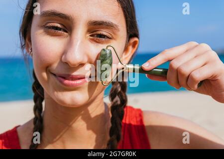 Content femelle utilisant le rouleau de jade et faisant le massage du visage tout en regardant la caméra sur la mer en été Banque D'Images