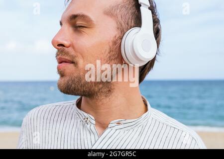 Jeune barbu insouciant dans une chemise élégante et décontractée, écoutant de la musique avec un casque sans fil et profitant d'une brise fraîche tout en passant une journée d'été Banque D'Images