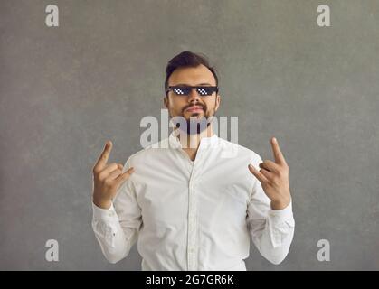 Portrait d'un jeune homme dans des lunettes de vie de voyou qui smirking et faisant le geste de signe de corne de rocher Banque D'Images