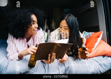 Des jeunes femmes gaies et multiraciales boivent de la bière et discutent de l'itinéraire de voyage avec la carte tout en se refroidissant ensemble dans une camionnette de camping pendant le voyage d'été Banque D'Images