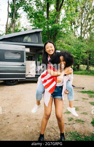 Pleine longueur de jeune femme afro-américaine joyeuse offrant un voyage de pigeyback au contenu de la petite amie asiatique avec drapeau américain tout en appréciant l'arrivée d'été Banque D'Images