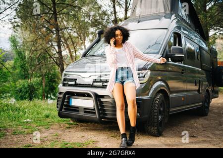 Une jeune femme afro-américaine sérieuse parle sur son smartphone et se gesticule tout en se penchant contre une voiture de camping garée dans la forêt en été Banque D'Images