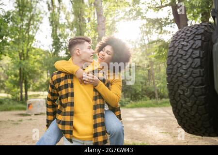 Jeune homme offrant une promenade en pigeyback à la petite amie noire optimiste vêtements décontractés tout en se tenant près de la voiture le jour ensoleillé dans la campagne Banque D'Images