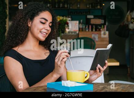 Jeune femme hispanique souriante aux cheveux maurires, en train de se reposer à table avec une tasse de café sur la terrasse du café Banque D'Images