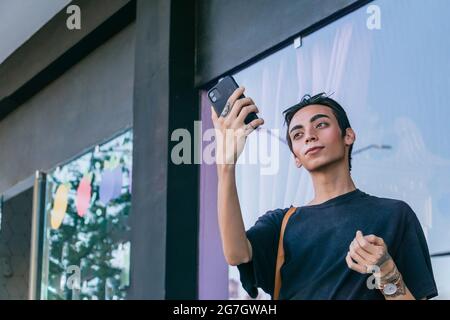 Faible angle d'un homme gay élégant debout près d'un bâtiment dans la rue et en train de prendre sa photo sur un smartphone Banque D'Images