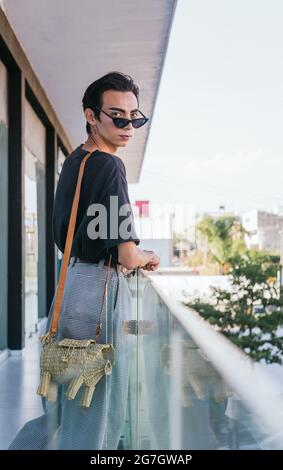 Vue latérale de gay masculin dans les lunettes de soleil tendance se pencher sur la rambarde de verre sur le balcon et vue loin Banque D'Images