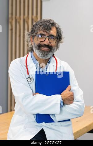 Joyeux homme âgé medic en uniforme et avec dossier debout dans la salle médicale dans la clinique et regardant la caméra Banque D'Images